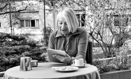 Foto de Mujer caucásica jubilada usando tableta relajándose en el jardín para desayunar, teniendo un videocall. Concepto de retiro, tecnología y relajación. - Imagen libre de derechos