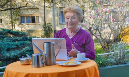 Foto de Mujer caucásica jubilada usando laptop relajándose en el jardín para desayunar, teniendo un videocall. Concepto de retiro, tecnología y relajación. - Imagen libre de derechos