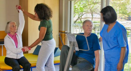 Foto de Concepto de rehabilitación. Ancianos jubilados pareja entrenamiento en un gimnasio supervisado por experto africano y asiático mujeres entrenadores. - Imagen libre de derechos