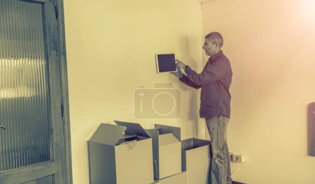 Photo for Moving to a new home with cardboard boxes on the floor. Smart home and technology concept, male hands pointing finger to a tablet with green screen integrated in house indoor wall - Royalty Free Image