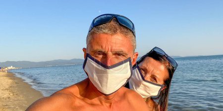 Foto de Hombre y mujer tomando selfie en la playa usando máscaras en tiempo covid - Imagen libre de derechos