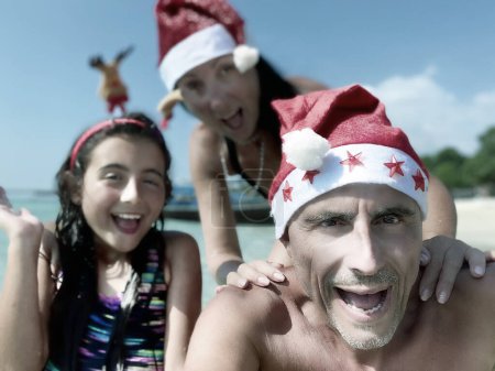 Photo for Family Tropical Christmas. Young girl together with her parents wearing Christmas Hats on a beautiful beach. - Royalty Free Image