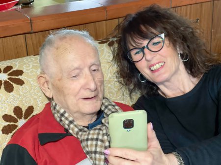Téléchargez les photos : Un homme âgé avec sa fille à la maison regardant le smartphone sur le canapé - en image libre de droit