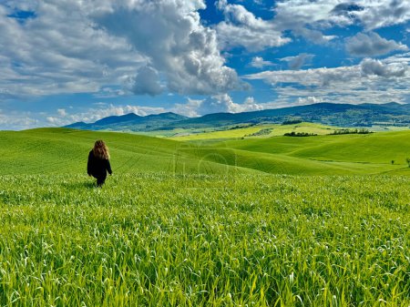 Photo for A woman walks through lush green fields under a clear blue sky, exuding tranquility and connection to nature. Rolling hills enhance the picturesque landscape, while her casual attire reflects a leisurely stroll. Predominant shades of green and blue c - Royalty Free Image