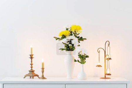 Photo for Chrysanthemums flowers in vases and burning candles on white interior - Royalty Free Image