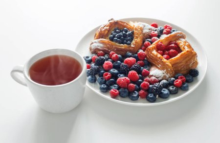 Téléchargez les photos : Danois aux framboises et aux bleuets avec une tasse de thé sur table blanche - en image libre de droit