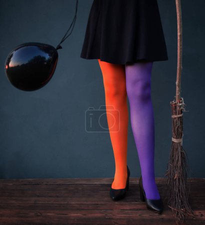 Téléchargez les photos : Jambes de sorcière en collants multicolores avec ballon noir et balai sur fond bleu - en image libre de droit