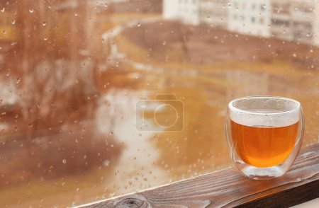 Téléchargez les photos : Thé chaud en verre thermique sur fond fenêtre d'automne avec gouttes de pluie - en image libre de droit