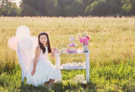 Téléchargez les photos : Heureuse petite fille en robe blanche avec des décorations d'anniversaire dans le champ d'été - en image libre de droit