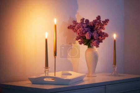 Foto de Flores lila en jarrón blanco y velas doradas sobre fondo pared blanca - Imagen libre de derechos