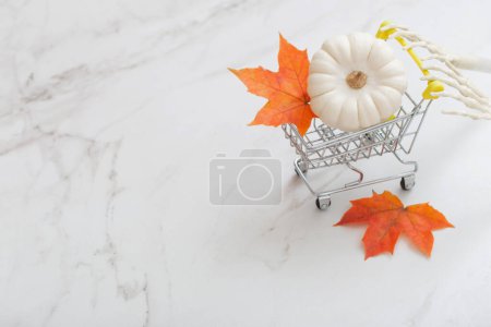 Téléchargez les photos : Citrouille blanche halloween dans un petit chariot d'épicerie avec des bras squelettes et des feuilles d'érable sur fond de marbre - en image libre de droit