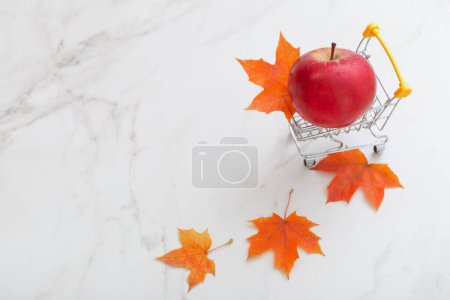 Foto de Manzana roja en un pequeño carrito de comestibles y hojas de arce sobre fondo de mármol blanco - Imagen libre de derechos