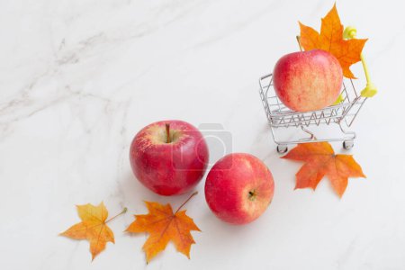Téléchargez les photos : Pomme rouge dans un petit chariot d'épicerie et feuilles d'érable sur fond de marbre blanc - en image libre de droit