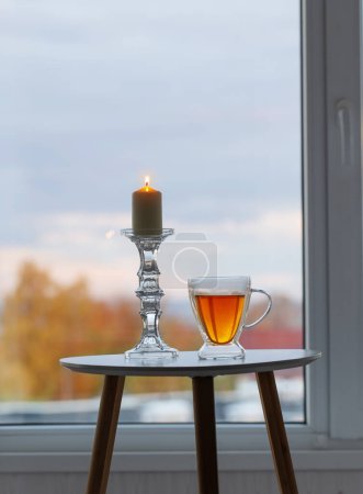Téléchargez les photos : Tasse thermique en verre avec thé et bougies allumées sur la fenêtre de fond avec paysage automnal - en image libre de droit