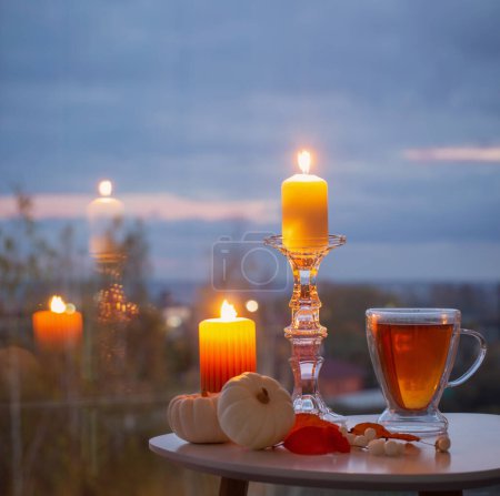 Téléchargez les photos : Tasse en verre avec thé et bougies allumées sur la fenêtre de fond avec paysage automnal - en image libre de droit