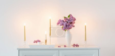 Téléchargez les photos : Fleurs lilas en vase blanc et bougies dorées sur fond blanc mur - en image libre de droit