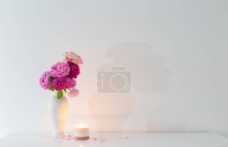 Foto de Rosas rosadas en jarrón y vela ardiente sobre fondo pared blanca - Imagen libre de derechos