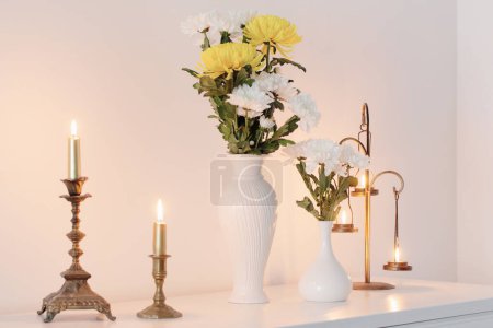 Photo for Chrysanthemums flowers in vases and burning candles on white interior - Royalty Free Image