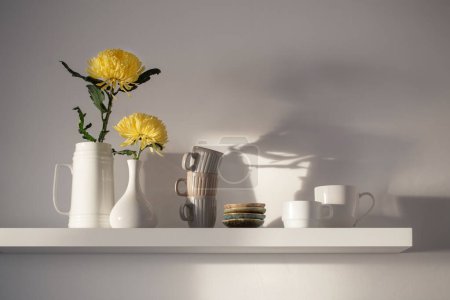 Téléchargez les photos : Chrysanthème jaune dans un vase et des tasses sur étagère blanche sur fond blanc - en image libre de droit