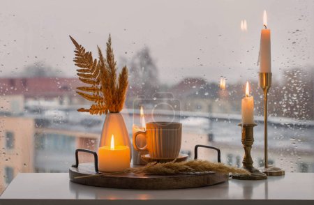 Foto de Naturaleza muerta de otoño con velas encendidas y una taza de café en el fondo de una ventana con gotas de lluvia - Imagen libre de derechos