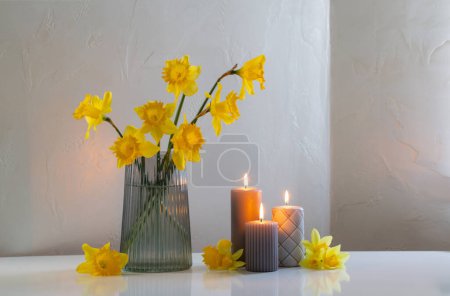 Téléchargez les photos : Jonquilles jaunes de printemps dans un vase en verre avec des bougies allumées sur fond blanc - en image libre de droit