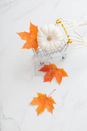 Foto de Calabaza de halloween blanco en pequeño carro de comestibles con brazos de esqueleto y hojas de arce sobre fondo de mármol - Imagen libre de derechos