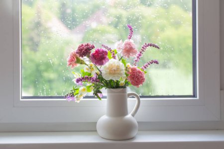 Foto de Rosa y púrpura flores de verano en jarra blanca en alféizar de ventana - Imagen libre de derechos