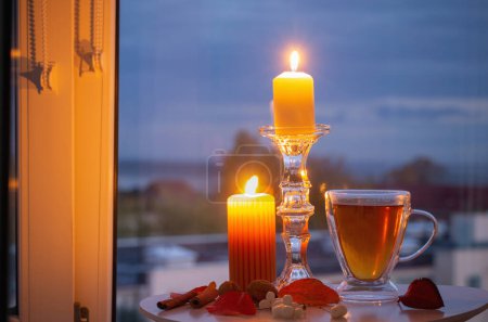 Foto de Taza de vidrio con té y velas encendidas en la ventana de fondo con paisaje otoñal - Imagen libre de derechos