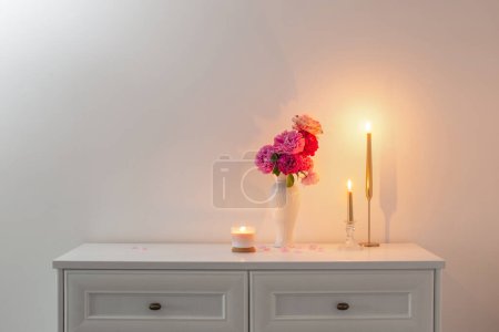 Foto de Rosas rosadas en jarrón y vela ardiente sobre fondo pared blanca - Imagen libre de derechos