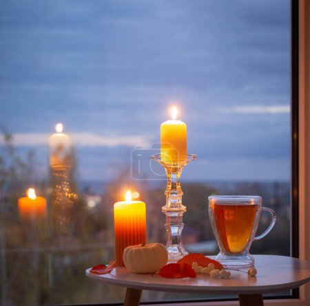 Téléchargez les photos : Tasse en verre avec thé et bougies allumées sur la fenêtre de fond avec paysage automnal - en image libre de droit