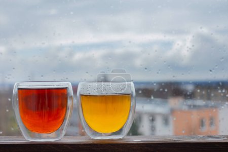 Téléchargez les photos : Thé chaud en verre thermique sur la fenêtre de fond avec des gouttes de pluie - en image libre de droit