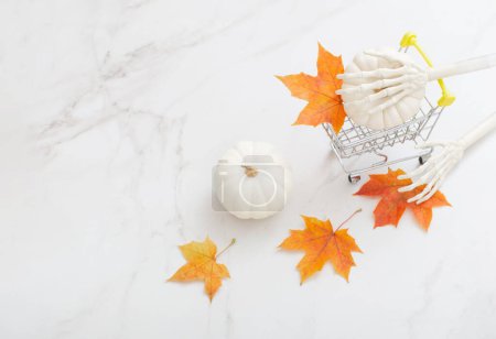 Photo for White halloween pumpkin in little grocery trolley with skeleton arms and maple leaves on marble background - Royalty Free Image