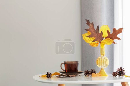 Photo for Cup of tea and autumn leaves in glass vase on background gray wall - Royalty Free Image