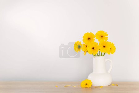Téléchargez les photos : Gerbera jaune dans une cruche blanche sur une étagère en bois sur le mur de fond - en image libre de droit