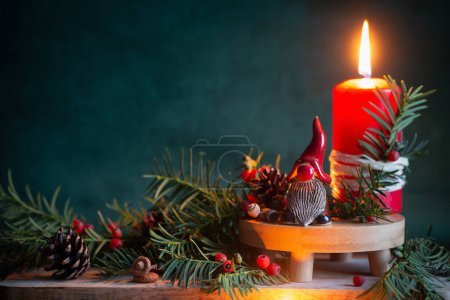 Foto de Vela ardiente roja con gnomo de cerámica en sombrero rojo con ramas de Navidad sobre fondo verde oscuro - Imagen libre de derechos