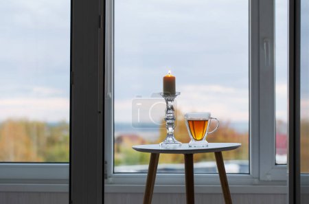 Téléchargez les photos : Tasse thermique en verre avec thé et bougies allumées sur la fenêtre de fond avec paysage automnal - en image libre de droit