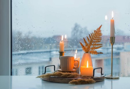 Téléchargez les photos : Nature morte d'automne avec des bougies allumées et une tasse de café sur le fond d'une fenêtre avec des gouttes de pluie - en image libre de droit