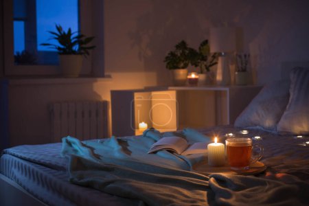 Photo for Cup of tea with burning candle on wooden tray on bed in bedroom in evening - Royalty Free Image