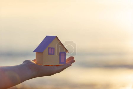 Téléchargez les photos : Une petite maison en papier à la main sur fond de mer au coucher du soleil, le concept de services immobiliers, un rêve d'une maison au bord de la mer - en image libre de droit
