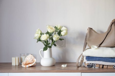 Photo for Knitted clothing and white roses in jug on modern dresser - Royalty Free Image