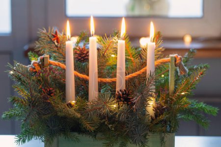 Photo for Advent decoration with fir branches and four burning candles in wooden basket on white table at home - Royalty Free Image