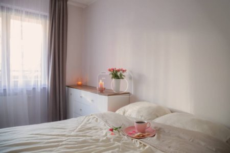 Téléchargez les photos : Tasse rose de café sur le lit avec des fleurs dans un vase dans une chambre blanche - en image libre de droit