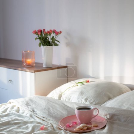 Téléchargez les photos : Tasse rose de café sur le lit avec des fleurs dans un vase dans une chambre blanche - en image libre de droit