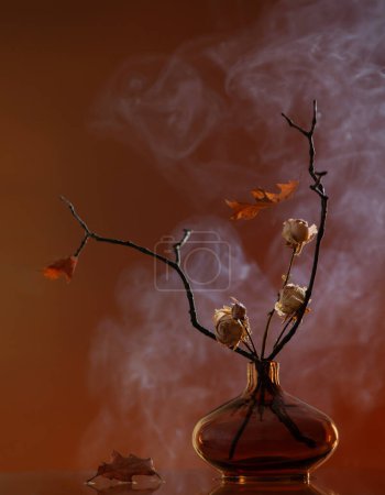 Foto de Bodegón de otoño con rosas secas y hojas de roble en jarrón de vidrio - Imagen libre de derechos