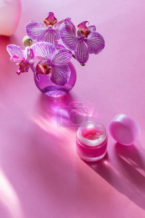 Téléchargez les photos : Baume à lèvres avec orchidée dans un vase en verre sur fond rose - en image libre de droit