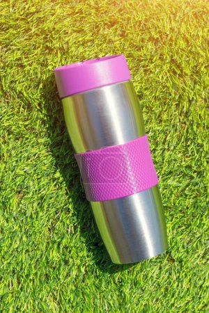 Photo for The stainless thermal flask on the green grass background with sunlight. The container for cold or hot beverages for outdoor activity. Selective focus. - Royalty Free Image
