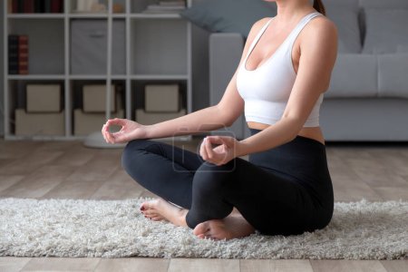 Foto de Mujer joven y deportiva practicando yoga en casa, sentada en pose de loto - Imagen libre de derechos