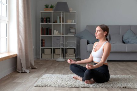 Foto de Mujer joven y deportiva practicando yoga en casa, sentada en pose de loto - Imagen libre de derechos