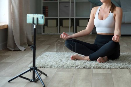 Foto de Mujer haciendo yoga en casa en la alfombra, sentada en posición de loto con su teléfono y trípode para grabar videos o transmisión en vivo para una clase de gimnasio en línea a través de una plataforma de redes sociales. - Imagen libre de derechos