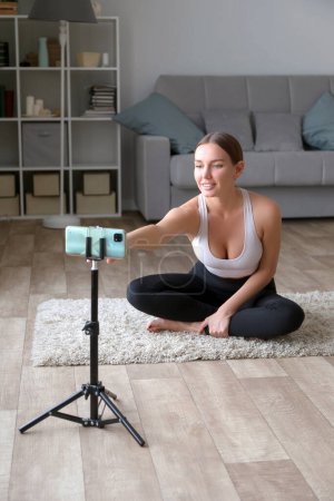 Téléchargez les photos : Femme assise sur le sol en pantalon de yoga enregistrant une vidéo de fitness en ligne avec son téléphone et son trépied, souriant à la caméra dans un cadre de salon - en image libre de droit